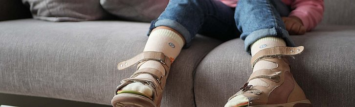 Kind sitzt auf Couch, im Vordergrund sind seine Füße mit Orthesen zu sehen