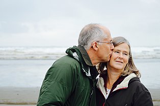 Ein Mann und eine Frau im mittleren Lebensalter stehen am Strand. Der Mann küsst die Frau auf die Wange.