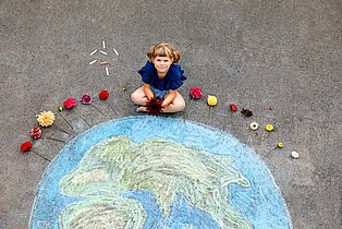 Die Erdkugel ist mit Kreide auf Asphalt gemalt. Davor sitzt ein Kind im Schneidersitz und blickt hinauf in die Kamera.
