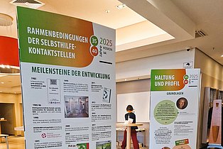 Zwei aufrecht stehende Informationssäulen mit Text und Grafiken in einem Innenraum, im Hintergrund eine Person an einem Stehtisch.