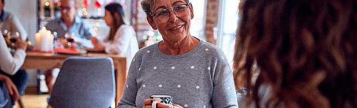 Person mit Kaffeetasse im Gespräch mit anderer Person, im Hintergrund weitere Personen an einem Tisch