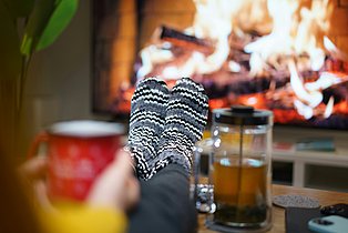 Ein Paar Füße in Wollsocken auf einem Couchtisch abgelegt. Tee und Kaminfeuer im Hintergrund.