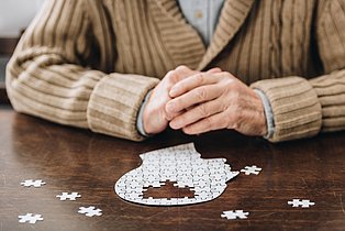 Ein Puzzle in Form eines Kopfes, oben in der Mitte fehlen einige Teile. Dahinter sind Hände und Strickjacke einer älteren Person zu sehen.